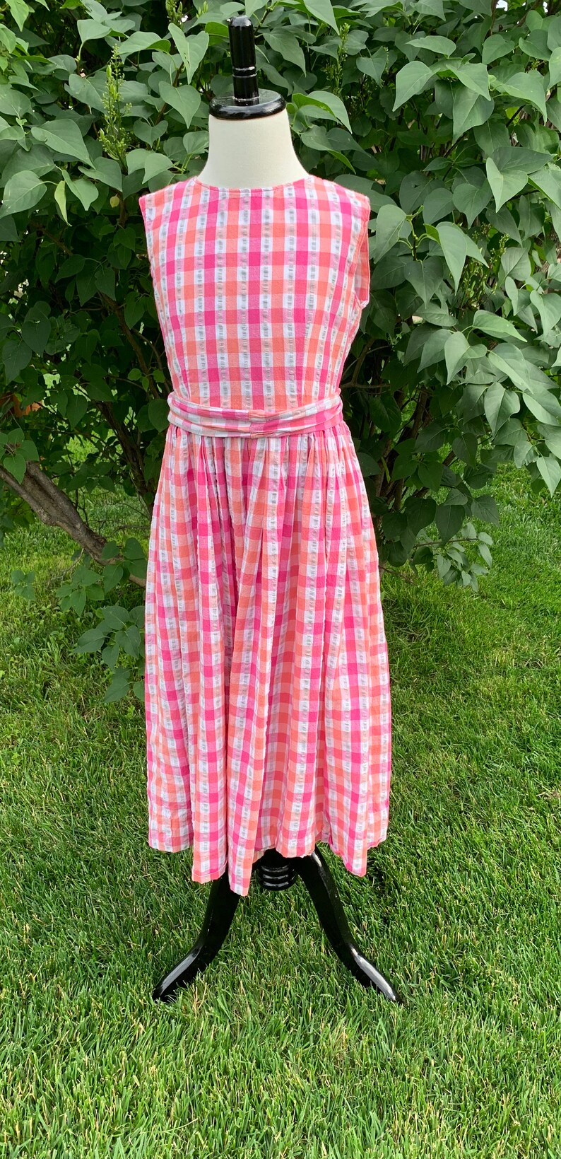 K&ouml;nnte beinhalten: Ein &auml;rmelloses Kleid in rosa und wei&szlig;em Gingham mit einem passenden G&uuml;rtel. Das Kleid befindet sich auf einer Puppe und wird vor einem gr&uuml;nen Gras Hintergrund pr&auml;sentiert.