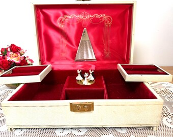 May include: A rectangular cream-coloured jewellery box with a gold-tone metal latch. The interior is lined with red velvet and features a triangular mirror, two side trays, and a rotating musical mechanism with three small dancing figurines.