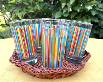 May include: Set of five clear drinking glasses with vertical stripes in various colors, including red, yellow, blue, and green. The glasses are arranged in a brown wicker basket, perfect for serving beverages.