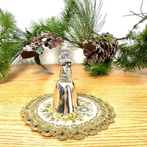 Pode incluir: Um sino de Natal prateado com uma figura do Pai Natal no topo, pousado sobre uma toalha de mesa bordada floral. O sino &eacute; rodeado por ramos de pinheiro, pinhas e bagas vermelhas, criando uma cena festiva.