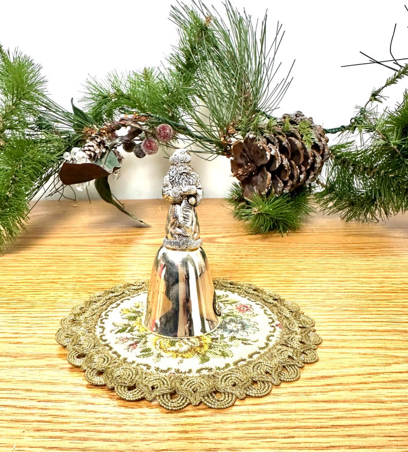 Pode incluir: Um sino de Natal prateado com uma figura do Pai Natal no topo, pousado sobre uma toalha de mesa bordada floral. O sino &eacute; rodeado por ramos de pinheiro, pinhas e bagas vermelhas, criando uma cena festiva.