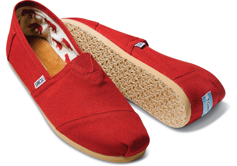 May include: A pair of red canvas slip-on shoes with a brown sole and the word "TOMS" on the side.