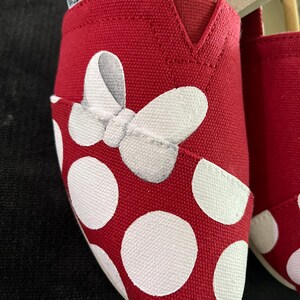 May include: Red canvas shoes with white polka dots and a white bow painted on the toe. The shoes are size 7.