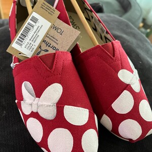 May include: Red canvas slip-on shoes with white polka dots and a white bow. The shoes have a TOMS label on the side.