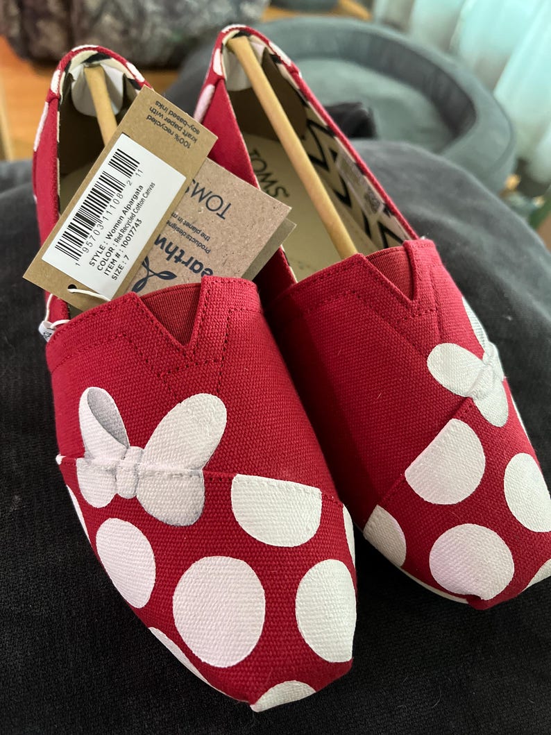 May include: Red canvas slip-on shoes with white polka dots and a white bow. The shoes have a TOMS label on the side.