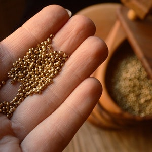 May include: A hand holding a pile of small, gold-coloured beads. The beads are round and smooth, and they are scattered in a loose pile.