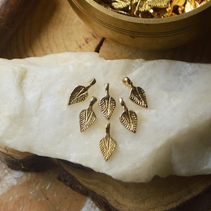 May include: A group of six gold leaf-shaped charms with a loop at the top. The charms are arranged in a cluster on a white surface.