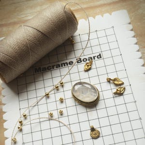 May include: A white gridded macrame board with a spool of beige cord, gold beads, and gold metal charms. The board has the text "Macrame Board" printed on it.