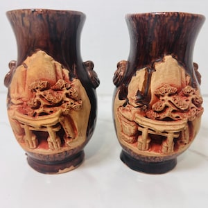 May include: A pair of dark brown ceramic vases featuring a raised, light tan relief design. The relief depicts a landscape with a torii gate, a pine tree, and rocky formations. Each vase has small decorative handles on the sides and a flared rim.