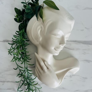May include: A white ceramic wall vase in the shape of a woman's head and shoulders, adorned with green faux foliage. The vase has a smooth, glossy finish and is mounted on a white marble-patterned surface.