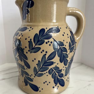 May include: A tan ceramic pitcher with a dark blue leaf and dot pattern. The pitcher has a curved handle and a spout for pouring. The design is hand-painted, giving it a rustic, farmhouse aesthetic.
