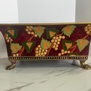 Könnte beinhalten: Ein dekorativer rechteckiger Behälter mit burgunderfarbenem Hintergrund, verziert mit gemalten Trauben und grünen Blättern. Der Behälter hat goldfarbene Verzierungen und steht auf vier goldfarbenen Klauenfüßen.