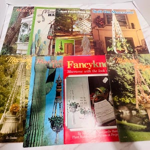 May include: A collection of vintage macrame books with colorful covers. Titles include "Macrame," "Soft On Macrame," and "Fancykno." The books feature images of macrame plant hangers and other crafts, with text such as "Macrame with the look."