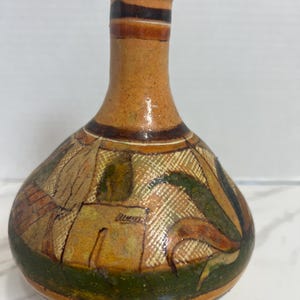 May include: A small, round, tan ceramic vase with a narrow neck. The body features hand-painted geometric patterns and stylized leaf motifs in shades of dark green, brown, and cream. A dark brown horizontal band encircles the neck and base.