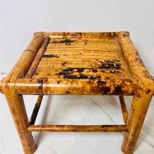 May include: A small square stool constructed from natural bamboo poles. The seat features a slatted design made of thin bamboo strips, while the frame consists of thicker, rounded bamboo legs and supports with a mottled brown and tan finish.