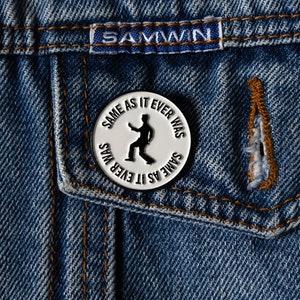 May include: A silver pin with the text "Same as it ever was" and a silhouette of a person dancing. The pin is on a blue denim jacket.