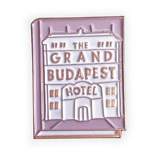 May include: A pink enamel pin featuring a white building with the text "The Grand Budapest Hotel" in gold lettering.
