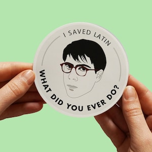 May include: A white round sticker with a black and white illustration of a person wearing glasses. The text on the sticker reads "I saved Latin. What did you ever do?"