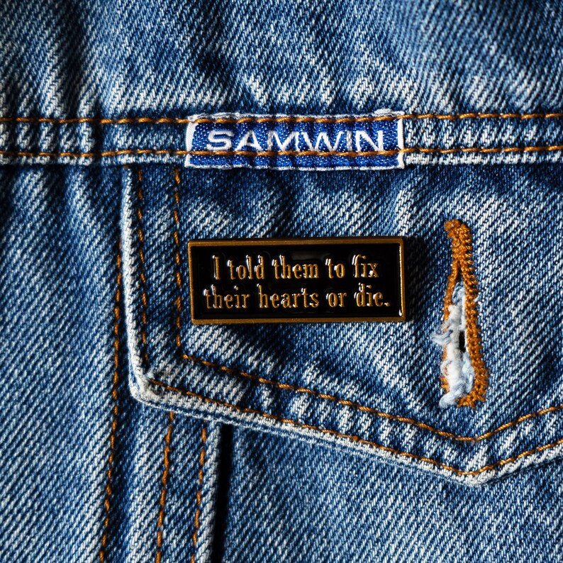May include: A black enamel pin with gold trim on a denim background. The pin says "I told them to fix their hearts or die."