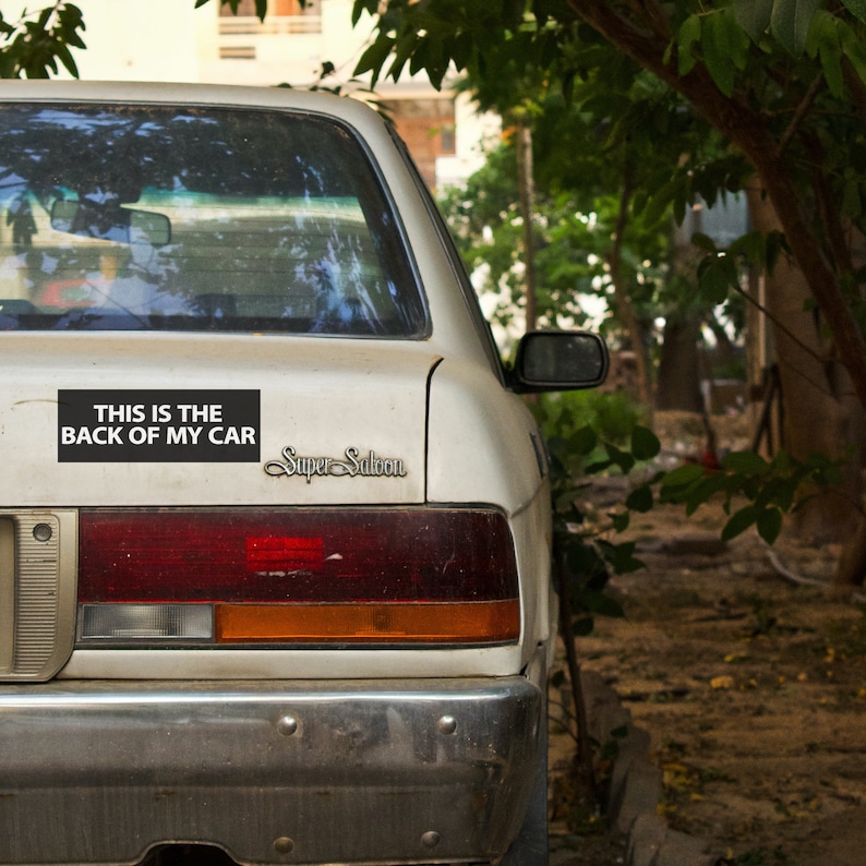 May include: A white car's rear with a bumper and taillights. A black rectangle sticker reads "THIS IS THE BACK OF MY CAR." The car has a "Super Saloon" emblem. The image is taken outdoors with trees in the background.