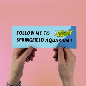 May include: A light blue sign with the text "FOLLOW ME TO SPRINGFIELD AQUARIUM!" and a cartoon fish swimming in blue water.