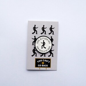 May include: White enamel pin with a black silhouette of a person dancing and the text "Same as it ever was" repeated around the edge. The pin is on a gray background with black silhouettes of people dancing. Below the pin is a black and white sign that says "You can't go back."
