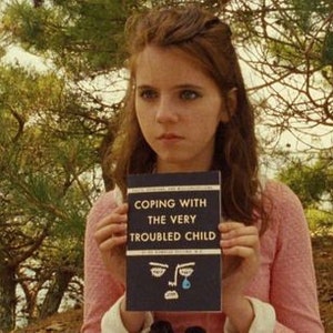 Pu&ograve; includere: Una giovane donna con lunghi capelli castani tiene in mano un libro intitolato "Coping with the Very Troubled Child". La copertina del libro presenta un viso di cartone animato blu con una singola lacrima.