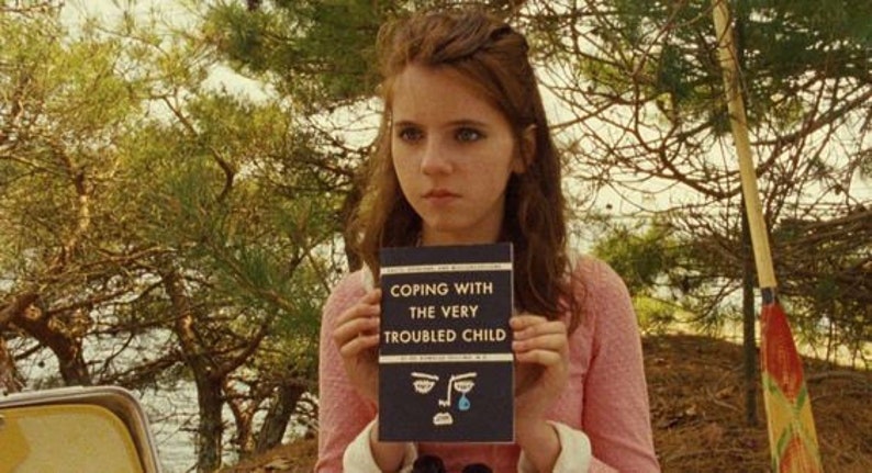 Pu&ograve; includere: Una giovane donna con lunghi capelli castani tiene in mano un libro intitolato "Coping with the Very Troubled Child". La copertina del libro presenta un viso di cartone animato blu con una singola lacrima.
