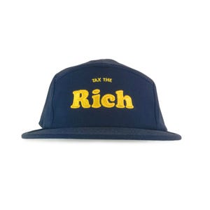 May include: Navy blue five-panel cap with a flat brim. The cap features the embroidered text "TAX THE Rich" in yellow, with the words stacked on top of each other. The hat is made of a soft material and is suitable for casual wear.