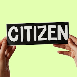 Könnte beinhalten: Ein schwarzer rechteckiger Aufkleber mit dem Wort "CITIZEN" in fetten weißen Buchstaben. Der Aufkleber wird zwischen zwei Händen vor einem hellgrünen Hintergrund gehalten.