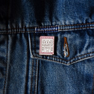 May include: A pink enamel pin featuring a building with the text "The Grand Budapest Hotel" in white.