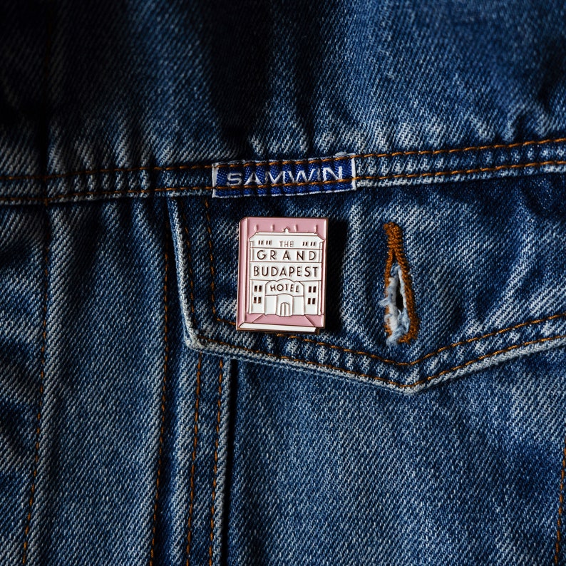 May include: A pink enamel pin featuring a building with the text "The Grand Budapest Hotel" in white.