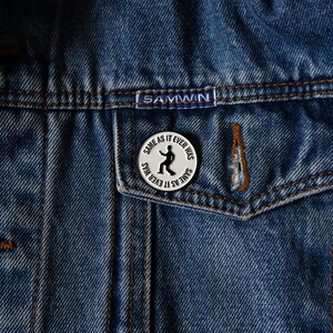 May include: A silver pin with a black silhouette of a person dancing and the text "SAME AS IT EVER WAS" repeated around the edge.