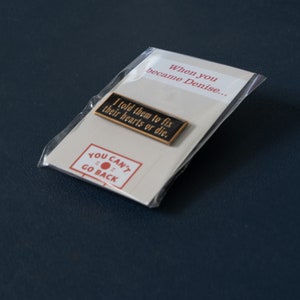 May include: A black and gold enamel pin with the text "I told them to fix their hearts or die."  The pin is packaged in a clear plastic bag with a white card that says "When you became Denise..." and "YOU CAN'T GO BACK" with a red circle.