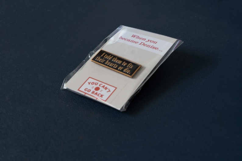 May include: A black and gold enamel pin with the text "I told them to fix their hearts or die."  The pin is packaged in a clear plastic bag with a white card that says "When you became Denise..." and "YOU CAN'T GO BACK" with a red circle.