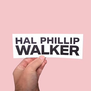 May include: A white rectangular sticker with the name "HAL PHILLIP WALKER" printed in bold black letters. The sticker is held by a hand against a light pink background.