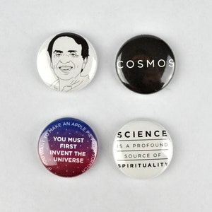 Puede incluir: Cuatro botones redondos con diferentes diseños. Un botón presenta una ilustración en blanco y negro de la cara de un hombre. Otro botón tiene la palabra "COSMOS" en texto blanco sobre fondo negro. Un tercer botón tiene un fondo degradado de azul y rojo con el texto "YOU MUST FIRST INVENT THE UNIVERSE". El cuarto botón tiene texto blanco sobre fondo blanco que dice "SCIENCE IS A PROFOUND SOURCE OF SPIRITUALITY".