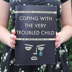 Pu&ograve; includere: Un libro nero con il titolo "Coping with the Very Troubled Child" di Dr. Romulus Trilling, M.D. La copertina presenta un'illustrazione in bianco e nero di un viso che piange.