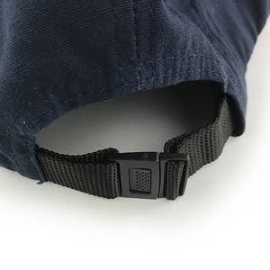 May include: Close-up of the back of a navy blue baseball cap. The cap features a black adjustable strap with a plastic buckle. The fabric of the cap is a solid navy blue color.