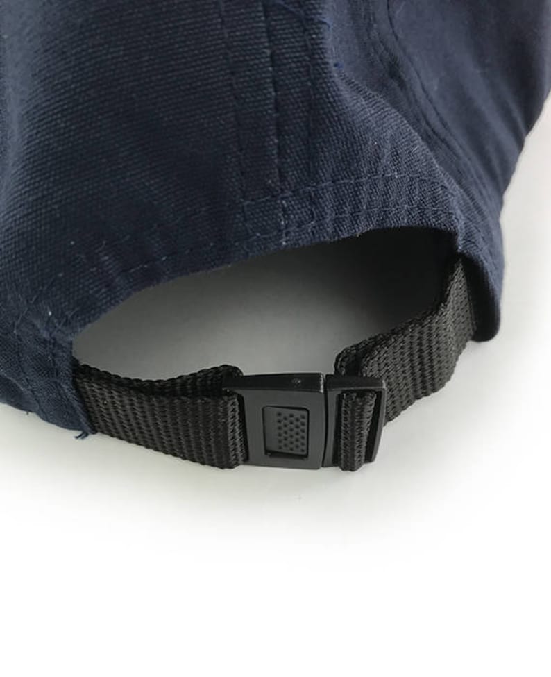 May include: Close-up of the back of a navy blue baseball cap. The cap features a black adjustable strap with a plastic buckle. The fabric of the cap is a solid navy blue color.