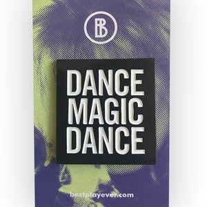 May include: Black enamel pin with white text that reads "DANCE MAGIC DANCE" on a white background. The pin is on a purple and yellow background with a white circle with the letter "B" inside.