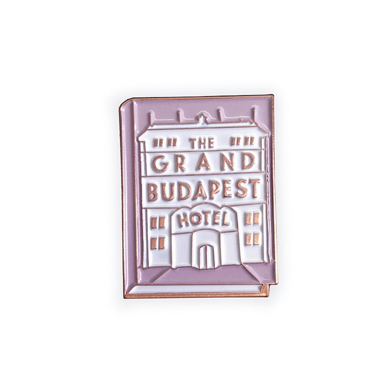 May include: A pink enamel pin featuring a white building with the text "The Grand Budapest Hotel" in gold lettering.