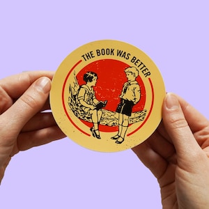 Puede incluir: Una pegatina circular amarilla con el texto "The Book Was Better" en negro. Dos niños están sentados en una hamaca roja, uno leyendo un libro.
