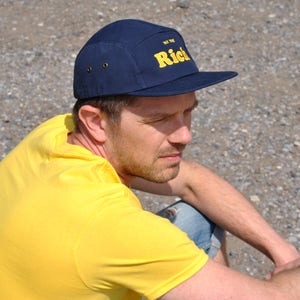 May include: Navy blue five-panel cap with a flat brim. The cap features the text "The Rich" in yellow block letters. The cap has two small metal eyelets on each side. The person in the image is wearing a yellow shirt.