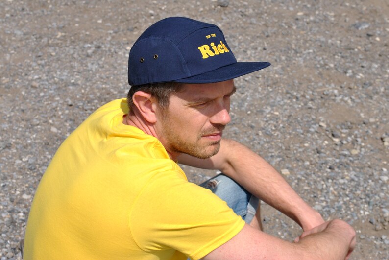 May include: Navy blue five-panel cap with a flat brim. The cap features the text "The Rich" in yellow block letters. The cap has two small metal eyelets on each side. The person in the image is wearing a yellow shirt.