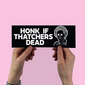 Puede incluir: Una pegatina para parachoques en blanco y negro con el texto "HONK IF THATCHERS DEAD" y una imagen de dibujos animados de Margaret Thatcher con una calavera como rostro.