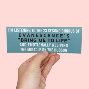 May include: A teal bumper sticker with white text is held up against a pink background. The text reads: "I'M LISTENING TO THE 23 SECOND CHORUS OF EVANESCENCE'S 'BRING ME TO LIFE' AND EMOTIONALLY RELIVING THE MIRACLE ON THE HUDSON."