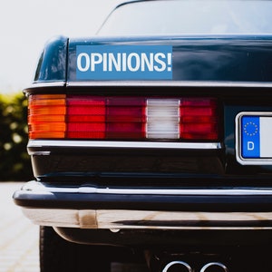 May include: A dark blue car with a blue sticker that says "OPINIONS!" on the back. The car has a European number plate with the letter "D" on it.