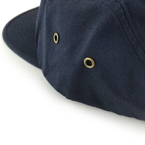 May include: Close-up of a navy blue canvas cap with a short brim. The cap features two brass-colored eyelets on the side for ventilation. The stitching is visible along the seams, and the cap is set against a white background.