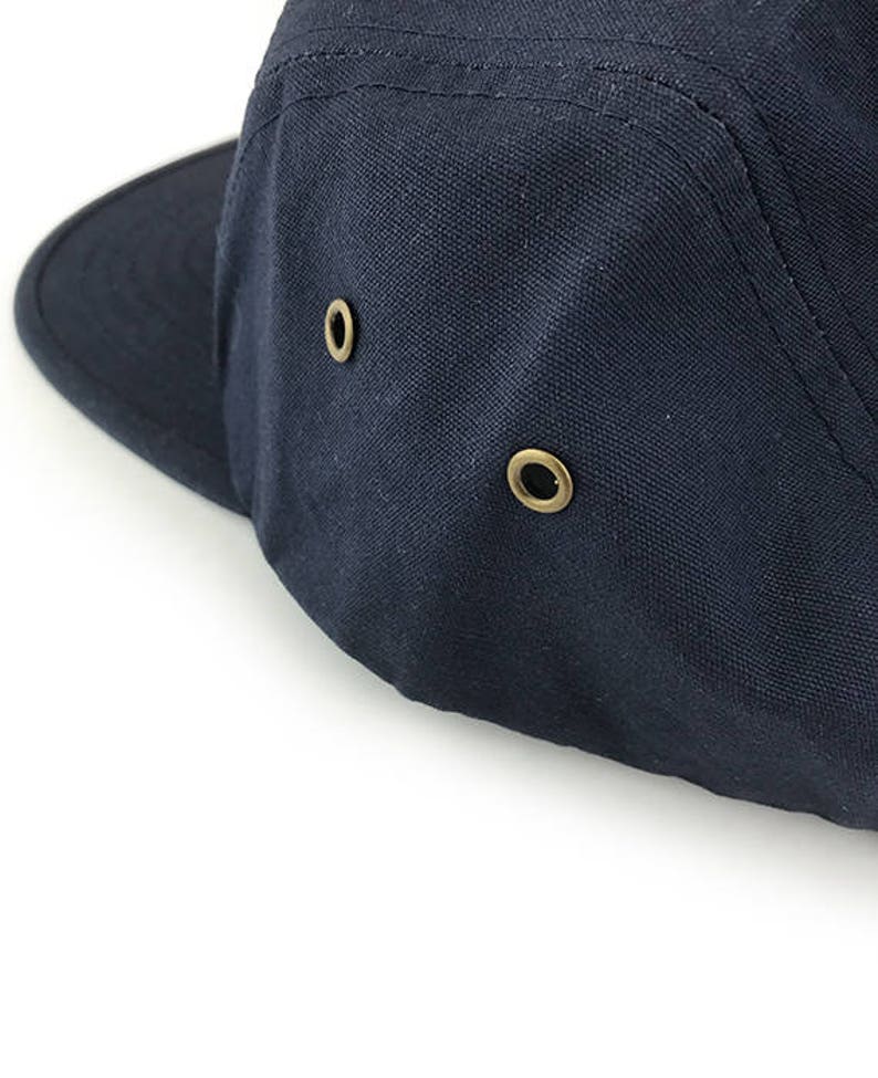May include: Close-up of a navy blue canvas cap with a short brim. The cap features two brass-colored eyelets on the side for ventilation. The stitching is visible along the seams, and the cap is set against a white background.